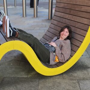 Gray is sitting in a yellow lounge chair outside of a London train station. They are smiling with the sun in their eyes
