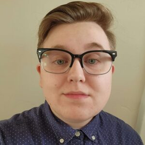 Skylar is smiling at the camera, he is wearing glasses and a navy button-up shirt with small white dots