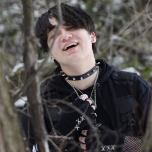 Tyler in mostly black clothing with copious amounts of jewelry laughing in with a branch covering his face a bit.