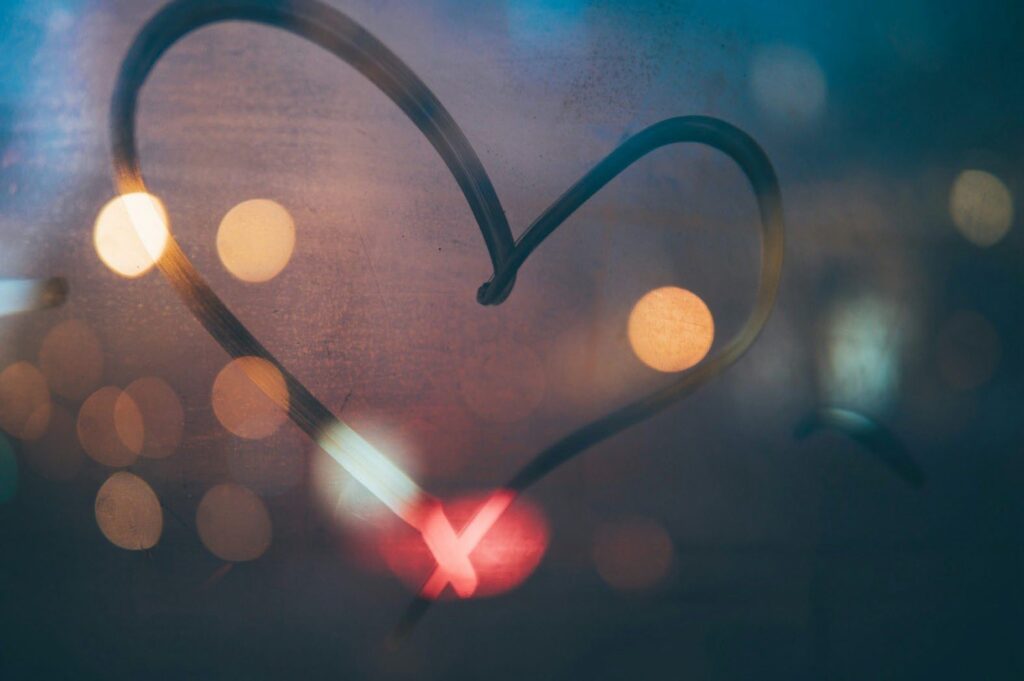 A close-up photograph of a heart shape drawn on a foggy or frosted glass surface. The heart is outlined with a dark, thin line. Through the glass, out-of-focus lights appear as blurred circles of white, orange, and red. The overall tone of the image is soft and muted, with a dreamy, romantic feel.