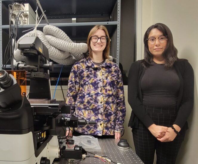 Two women students stand near science equipment.