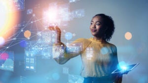 Person with black curly hair, yellow shirt, stands in front of digital see through board working on programming