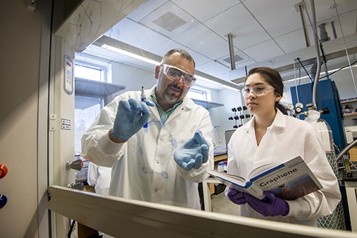 Dr. David Estrada and a female student in white lab coats and saftey glasses review equations written on glass inside a lab facilty