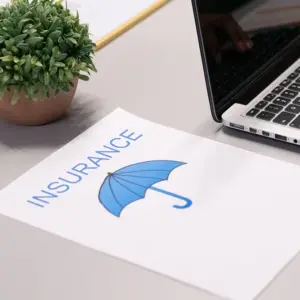 paper with the word "INSURANCE" and an umbrella symbol on it, lying on a desk with a laptop