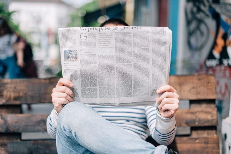 Person sitting on a bench outdoors, face hidden by a newspaper they're reading, in a casual striped outfit.