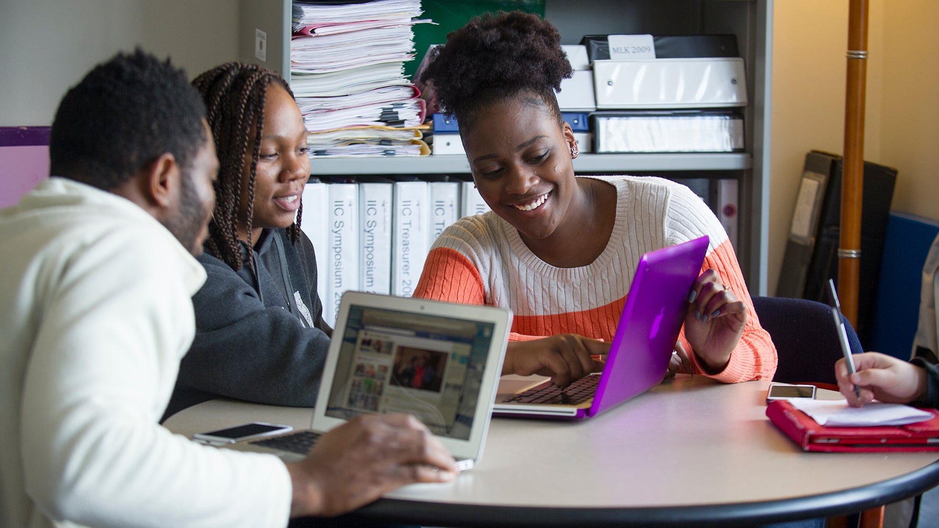 Three students sitting at a table talking and smiling looking at two laptop computers