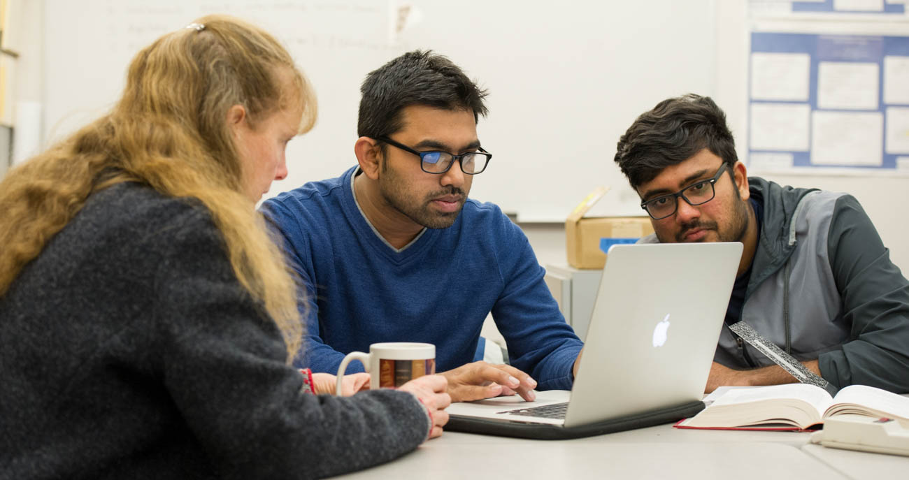 A professor sitting at a desk looking at a computer and talking to two students on either side of him