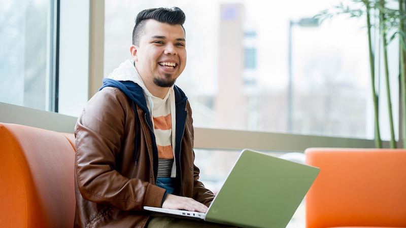 Student in a brown jacket using a laptop on an orange couch in a bright room with large windows.