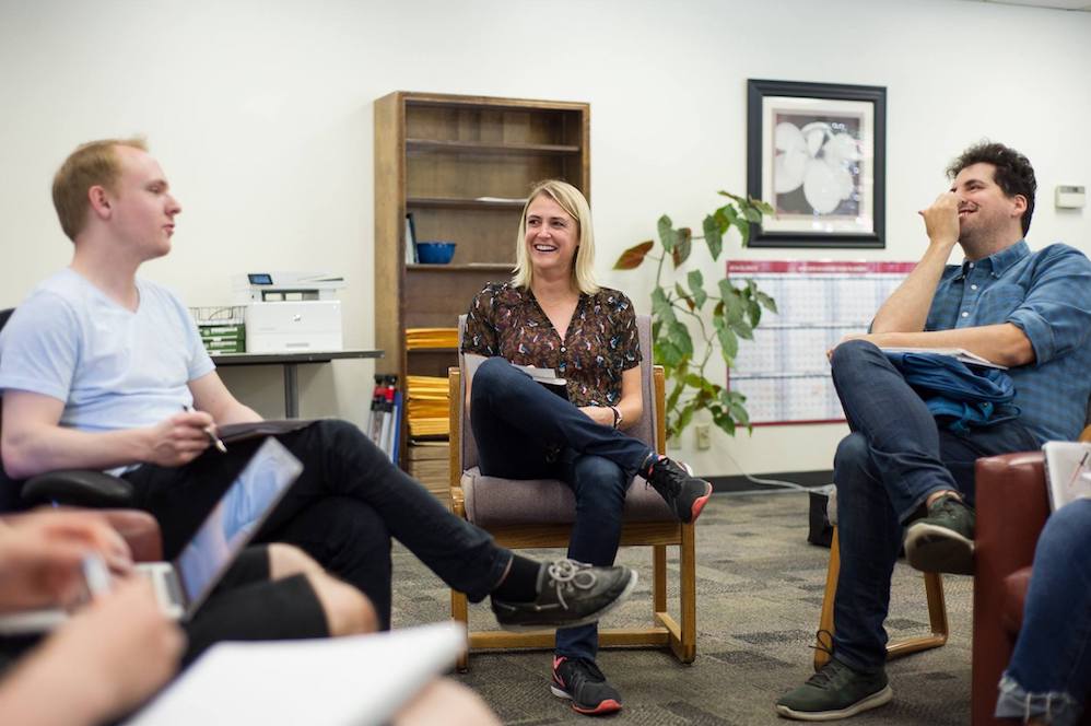 A blond woman sits between two men, all of them laughing and holding printed pages