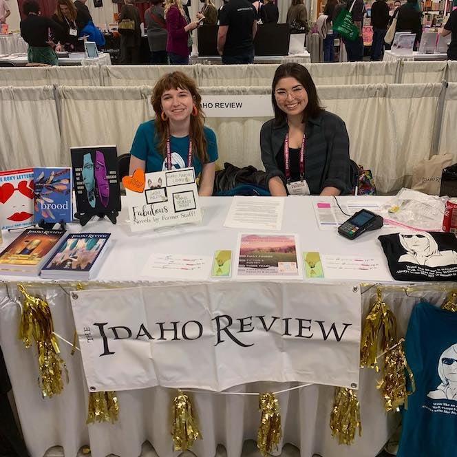 Two women sit and smile behind a booth with a sign that reads "Idaho Review" with people in the background.