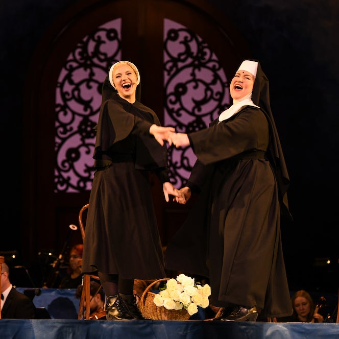 Two actors in nun costumes hold hands against a gothic backdrop.