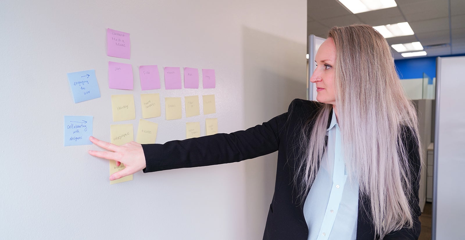 Jessi boyer in white shirt and black jacket points to sticky notes on a white board