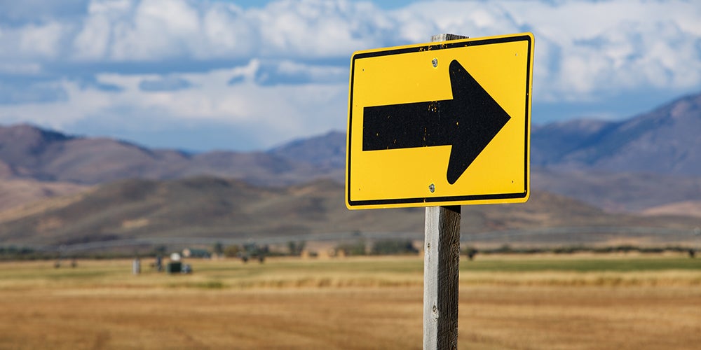A yellow road sign in a rural area with a large black arrow pointing right
